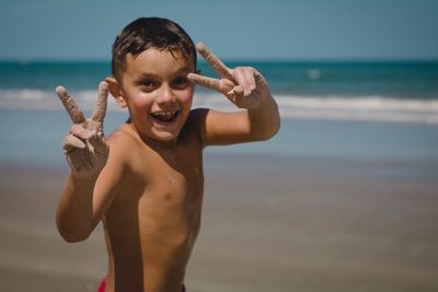 Portrait of happy shirtless boy showing peace sign while standing at beach