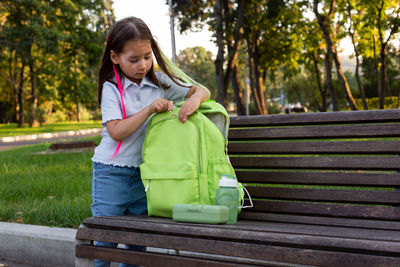 Young girl organizing backpack on park bench amidst greenery