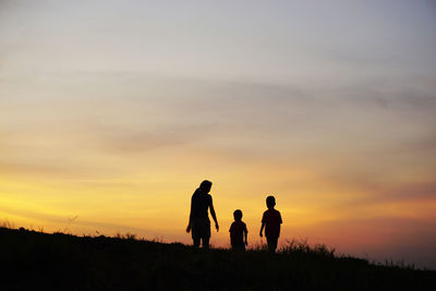 Silhouette mother walking with sons on field against orange sky