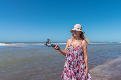Woman with a hat standing on the beach while filming with a mobile attached on a gimbal stabilizer