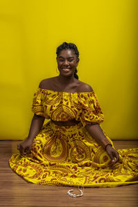 Young beautiful woman sitting on wooden floor, wearing colorful dress. against yellow background.