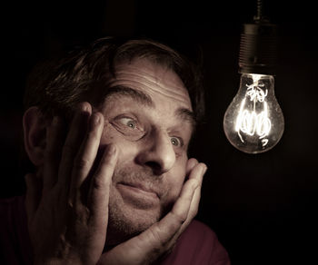 Close-up of smiling mature man looking at illuminated light bulb in darkroom