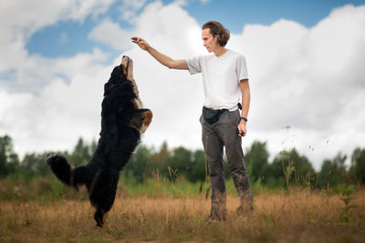 Man training dog on grassy land against cloudy sky