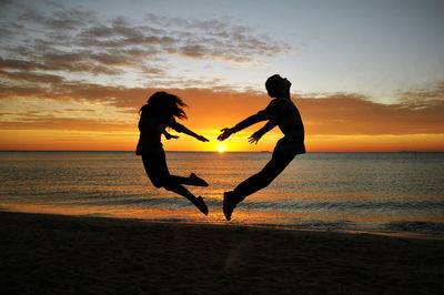 Full length of man and woman jumping at beach against sky during sunset