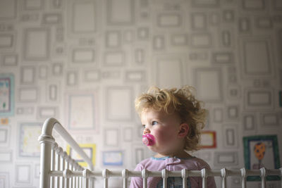 Low angle view of cute girl with pacifier in mouth looking away while standing against wall at home