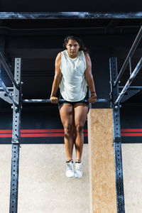 Full body of strong hispanic female athlete in sportswear doing exercise on metal crossbar during intense workout in light gym