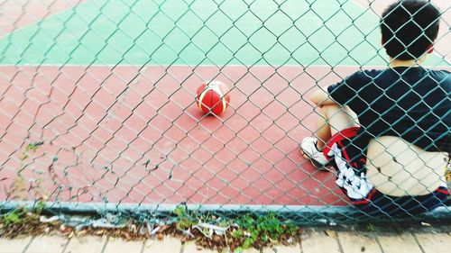 Rear view of boy sitting in front of basket ball on court