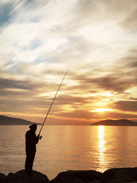 Silhouette man fishing in sea against sunset sky