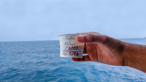 Close-up of hand holding coffee and tea by sea against sky. man enjoying coffee and tea