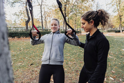 Happy friends exercising with straps while standing by tree