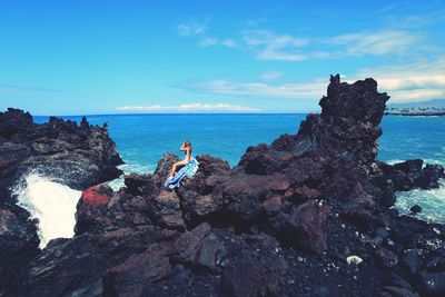 Side view of young woman sitting rock formations by sea against blue sky