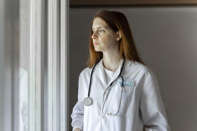 Thoughtful young female doctor looking through window while standing at home office