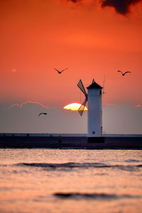 Silhouette birds flying over sea against orange sky and lighthouse in shape of a mill