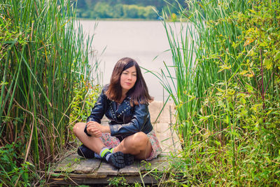 A lovely young woman with black hair is sitting in a thicket of tall grass on the river bank.