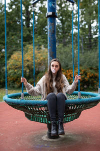 Young woman blowing bubble while sitting on outdoor play equipment