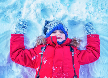 Smiling caucasian boy in red jacket and blue knit cap is lying on the snow. portrait. close-up.