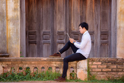 Side view of young man using digital tablet while sitting on retaining wall against building