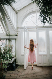 Back view of unrecognizable female with long ginger hair opening door of greenhouse with plants