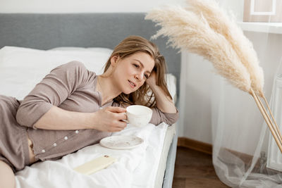 Young beautiful woman in light pajamas enjoys morning in bed with coffee in cozy bedroom.
