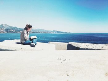 Side view of young woman reading book on retaining wall by sea against sky