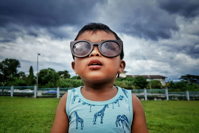 Close-up of boy wearing sunglasses while standing on field against cloudy sky