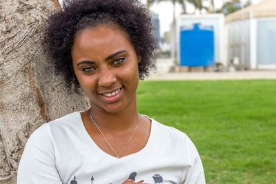 Portrait of smiling young woman standing by tree at park