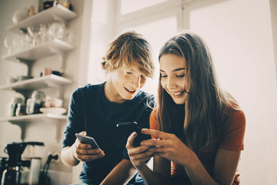 Close-up of friends using smart phone while sitting at kitchen counter against window