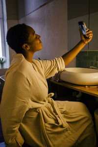 Smiling woman taking selfie through smart phone while sitting in bathroom