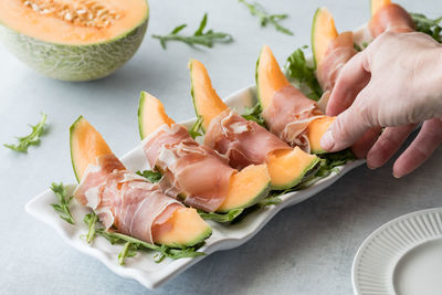 A hand reaching for a cantaloupe slice that is wrapped in prosciutto.