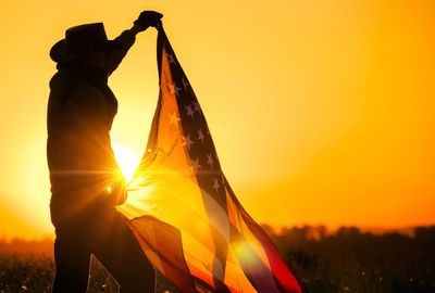 Silhouette man holding american flag on field against clear orange sky