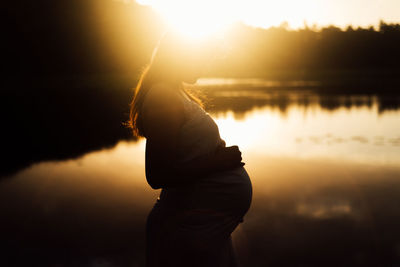 Side view of woman standing by lake against sky during sunset