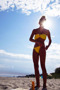 Full length of woman applying suntan lotion while standing at beach against sky