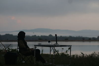 Man sitting on seat by lake against sky during sunset