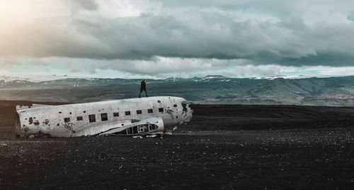 Woman standing on abandoned airplane at beach against cloudy sky