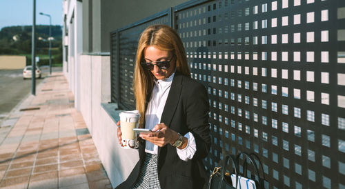 Businesswoman using smart phone holding coffee cup outdoors