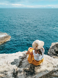 Young woman in yellow skirt sitting on edge of cliff above sea.