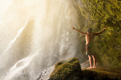 Full length of shirtless young man standing on  a cave inside a waterfall.