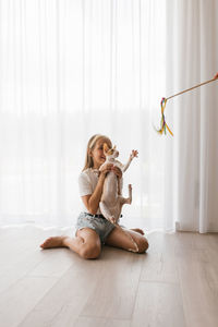 Caucasian teen girl sitting on the floor playing with a cat cornish rex with a stick teaser