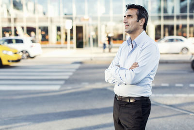 Businessman with arms crossed looking away while standing by city street