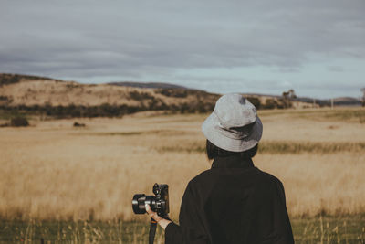 Rear view of woman wearing hat standing with camera on field against sky