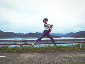 Full length of young woman jumping on field by lake against sky