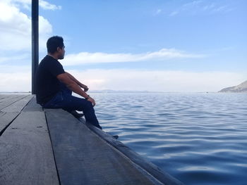 Side view of man sitting on wooden pier at sea against sky