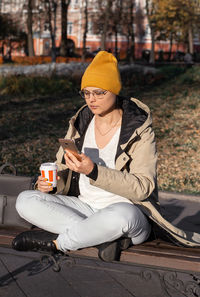  woman with a cup of drink sits cross-legged and looks thoughtfully into smartphone. street portrait