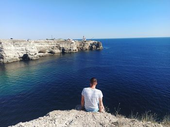 Rear view of man sitting on rock by sea against sky