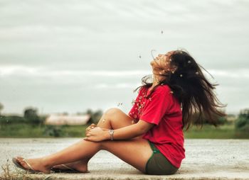 Side view of young woman with flowers sitting on road against cloudy sky