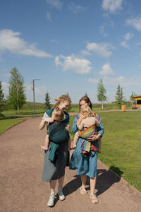 Full length of smiling caucasian mothers carrying their babies in slings at park on sunny day