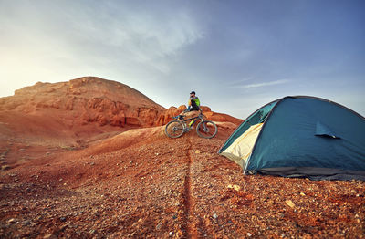 Mid distance view of hiker sitting by mountain bike on rocks at desert with tent in foreground