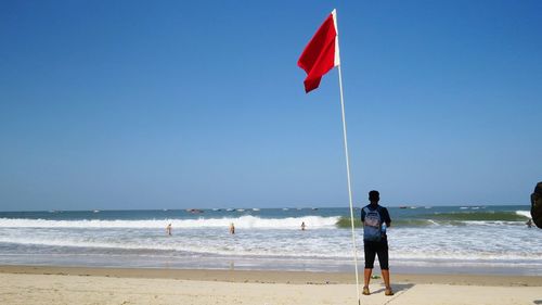 Rear view of man standing by flag on shore at beach against clear blue sky