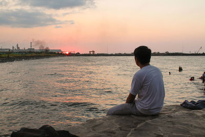 Rear view of man sitting on beach against sky during sunset