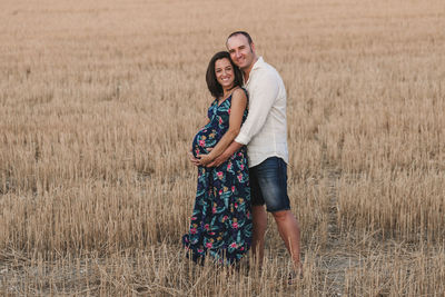 Portrait of smiling man touching pregnant girlfriend belly while standing on field